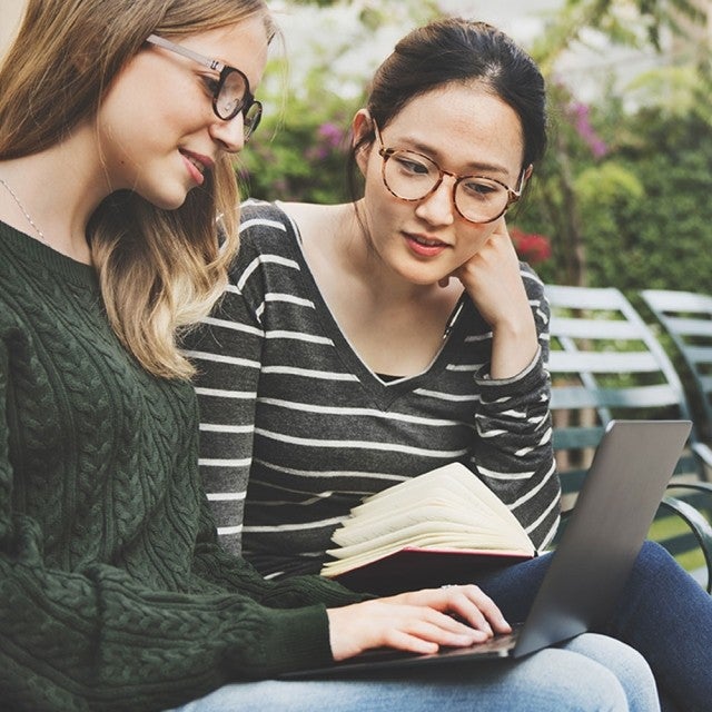 Two students sitting on park bench working on laptop